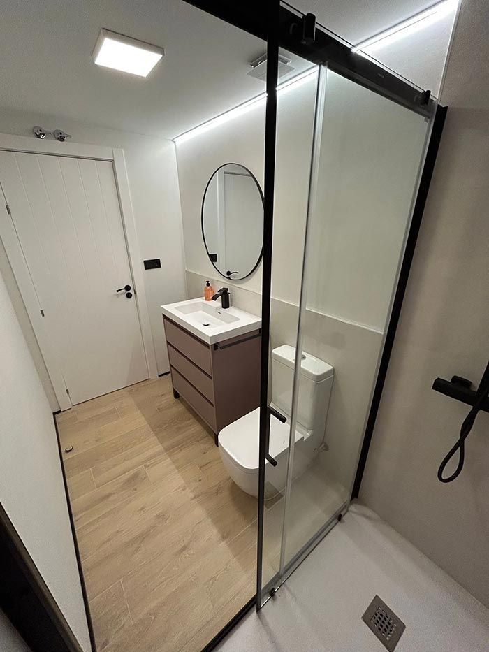 Pequeño baño moderno con ducha de cristal, tocador e inodoro; piso con apariencia de madera clara.