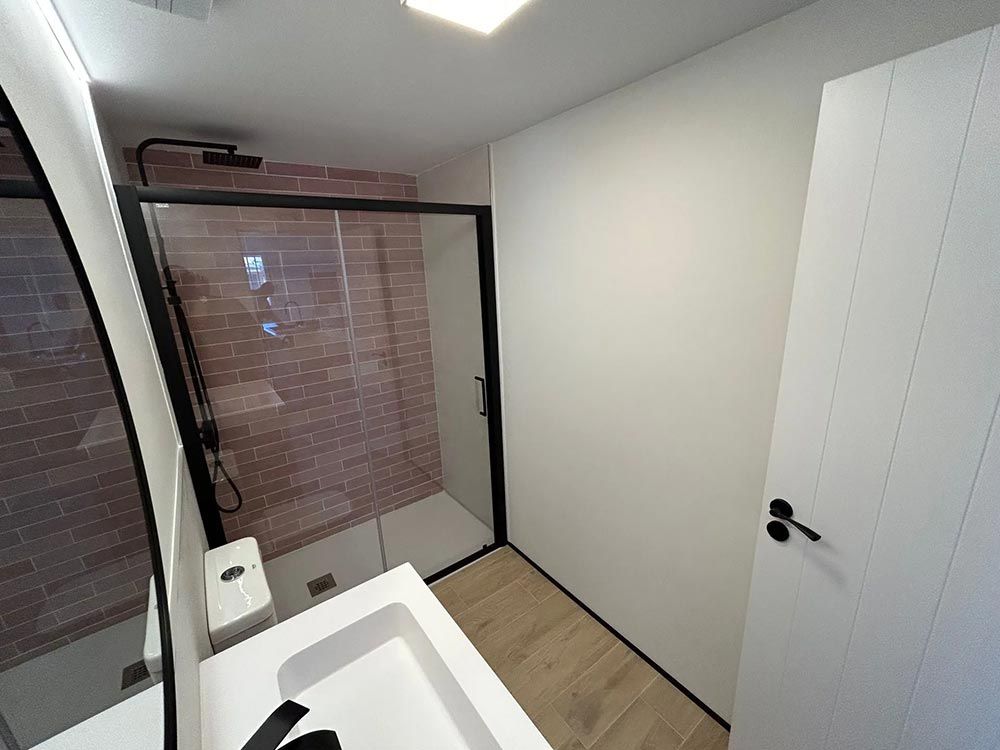 Baño moderno con ducha de ladrillo, marco negro, lavabo blanco, puerta y suelo de madera clara.