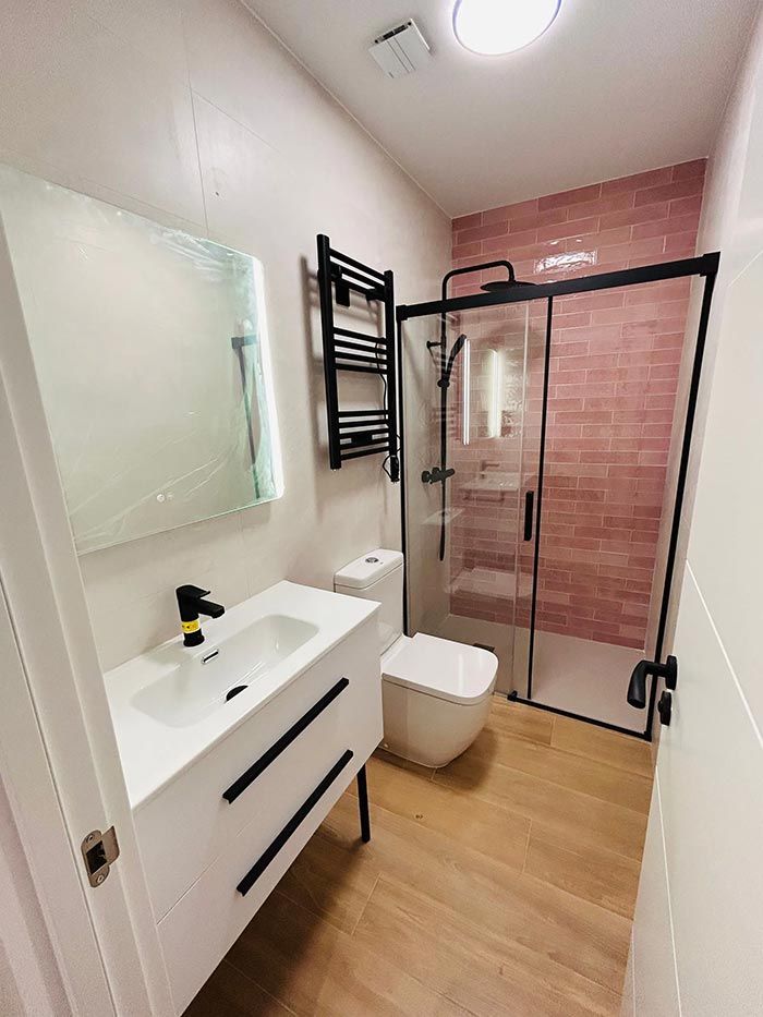 Baño moderno con tocador blanco, detalles en negro, ducha de vidrio, pared de ladrillo rosa y piso con apariencia de madera.