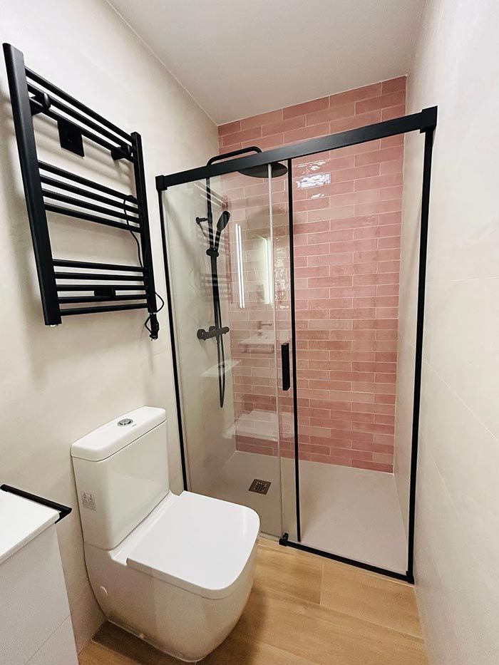Baño moderno con ducha de azulejos rosas, puerta de vidrio con marco negro, inodoro y toallero.