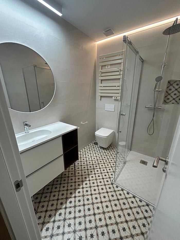 Baño moderno con espejo redondo, tocador blanco, suelo estampado y ducha de cristal.