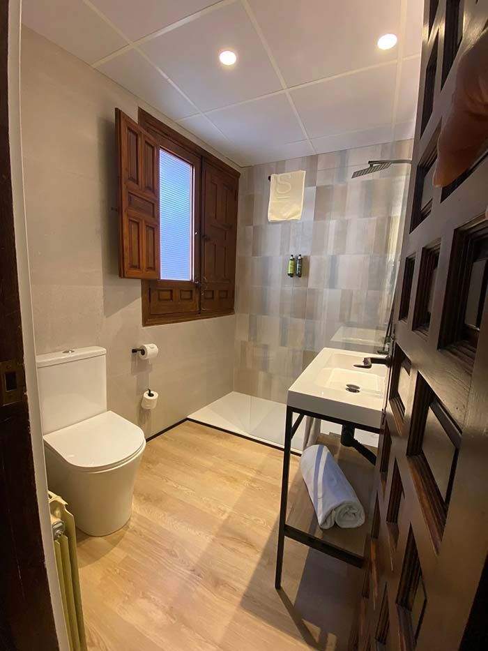 Baño con inodoro, ducha, lavabo y ventana con contraventanas de madera.