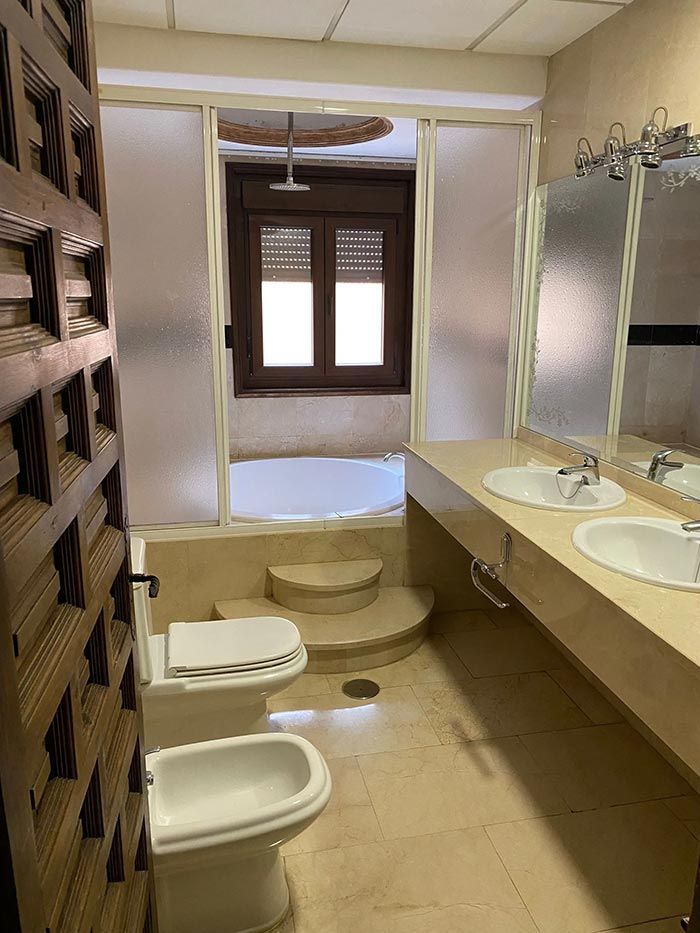 Baño con bañera, lavabo doble, inodoro y bidé. Incluye azulejos claros y una puerta decorativa de madera.