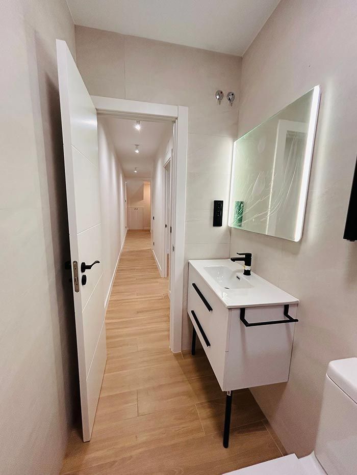 Baño estrecho con tocador blanco, espejo y puerta abierta a un pasillo con puertas e iluminación empotrada.