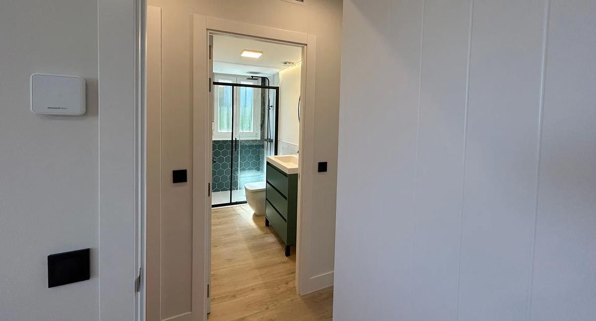 Pasillo que da acceso a un baño. Paredes y marcos de puertas blancos. Tocador verde y puerta de ducha negra visibles.