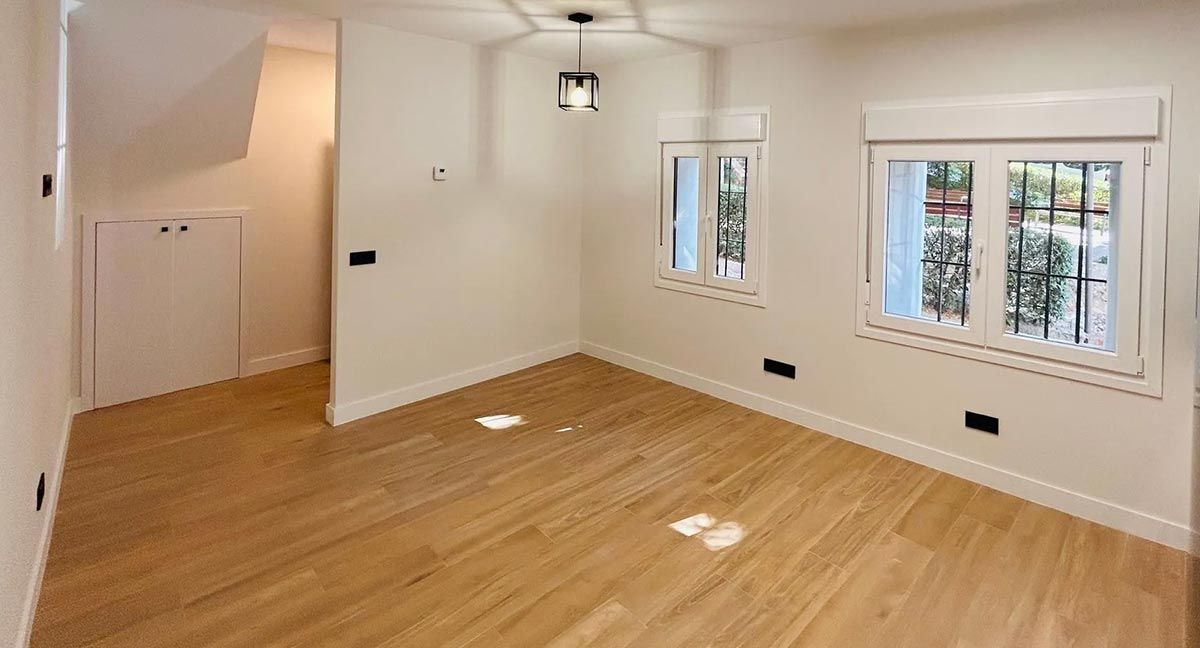 Habitación vacía con piso de madera clara, paredes blancas, dos ventanas y una lámpara.