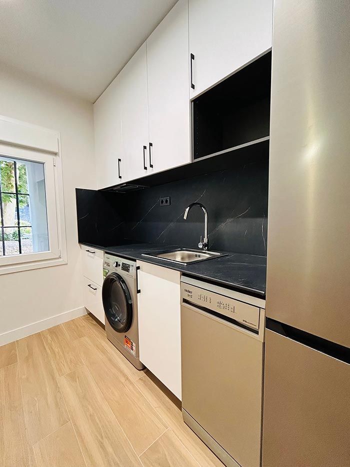 Lavandería con gabinetes blancos, protector contra salpicaduras negro, electrodomésticos de acero inoxidable y una ventana.