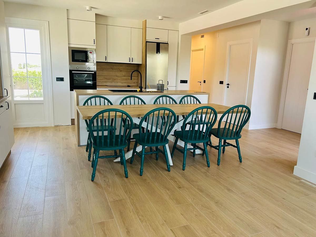 Cocina luminosa con una gran mesa de madera y sillas color verde azulado. Armarios blancos, suelos de madera y una ventana.