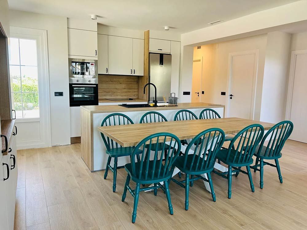 Cocina moderna con gabinetes blancos, isla de madera, sillas color verde azulado y electrodomésticos de acero inoxidable.