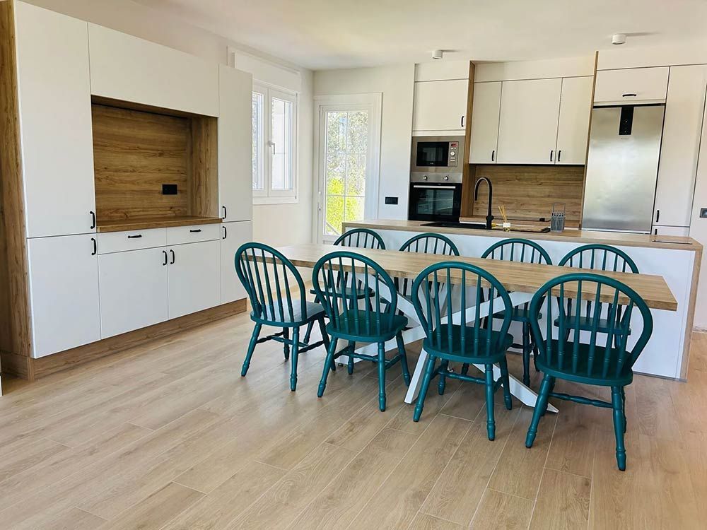 Cocina con gabinetes blancos, detalles de madera, sillas verde azulado alrededor de una mesa de madera clara y electrodomésticos.