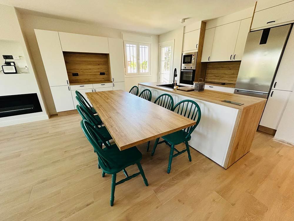 Cocina moderna con isla de madera, mesa larga de madera y sillas verdes. Muebles blancos y electrodomésticos de acero inoxidable.