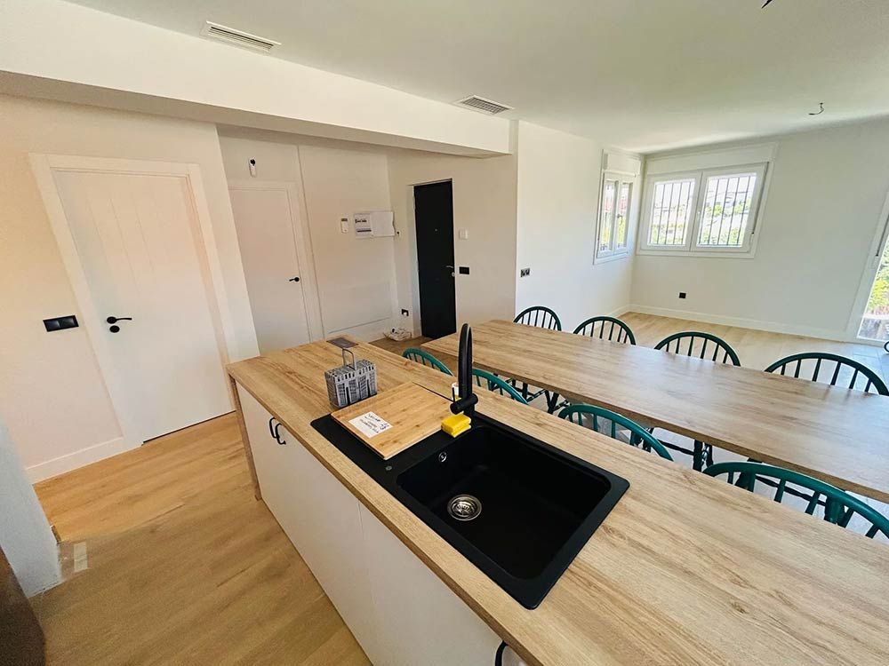 Cocina moderna con isla, fregadero y mesa de comedor larga. Madera clara y detalles en blanco, fregadero y grifo negros.