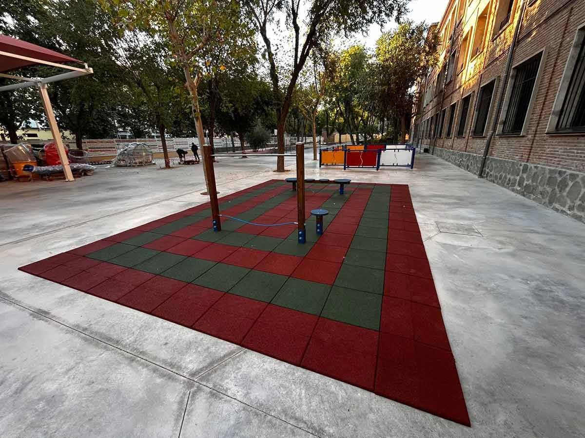 Parque infantil con baldosas de caucho rojas y verdes, árboles y equipos de juego contra un edificio.