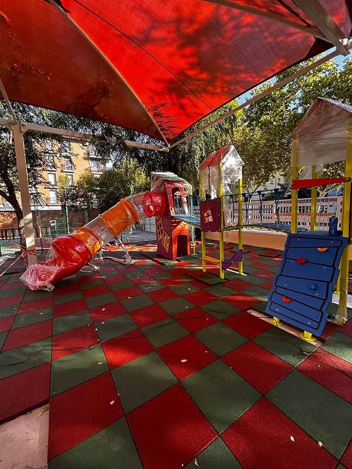 Parque infantil con tobogán rojo y naranja, pared de escalada azul, suelo a cuadros rojos y verdes, cubierto por una cortina roja.