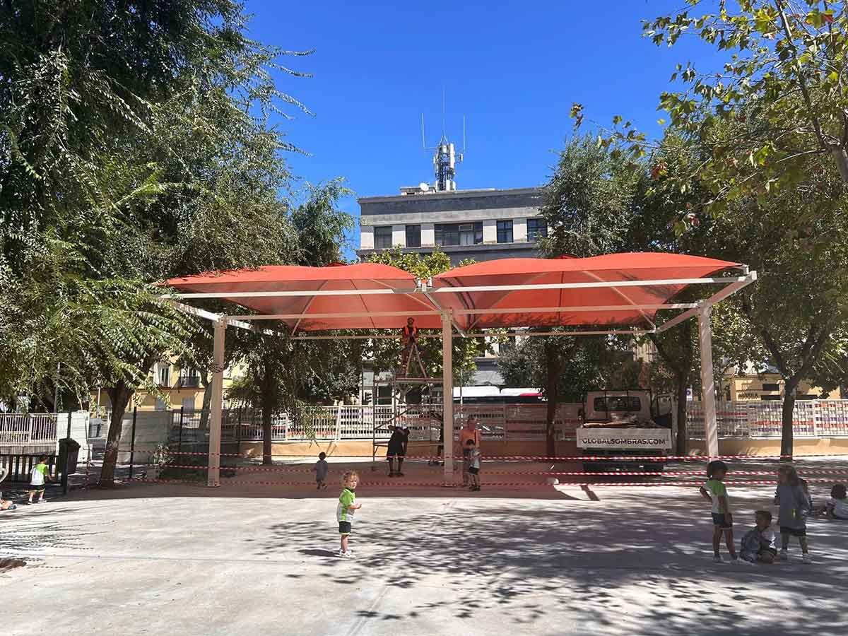 Dos estructuras de sombra con techo rojo en un parque. Niños juegan en el suelo de hormigón, árboles rodean el área y un edificio al fondo.