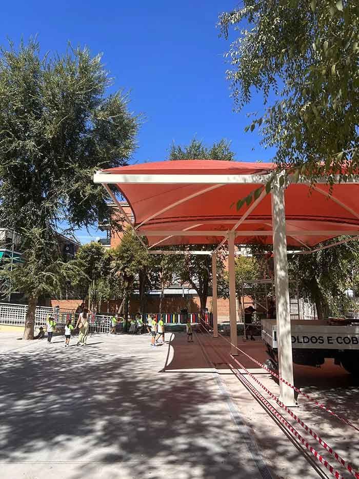 Un dosel rojo da sombra a un grupo de personas, bajo un cielo azul brillante.