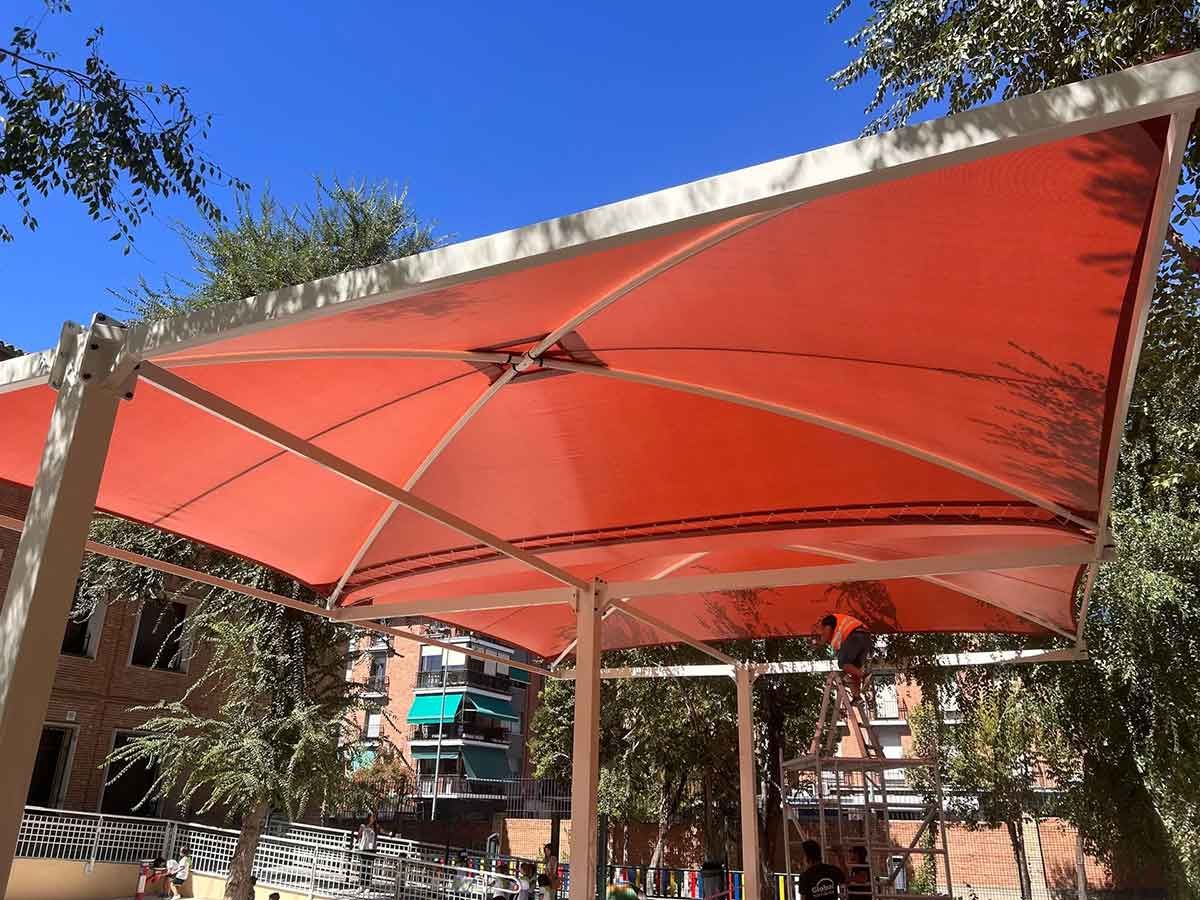 Estructura de sombra naranja sobre un espacio público con una persona en una escalera. Cielo azul de fondo.