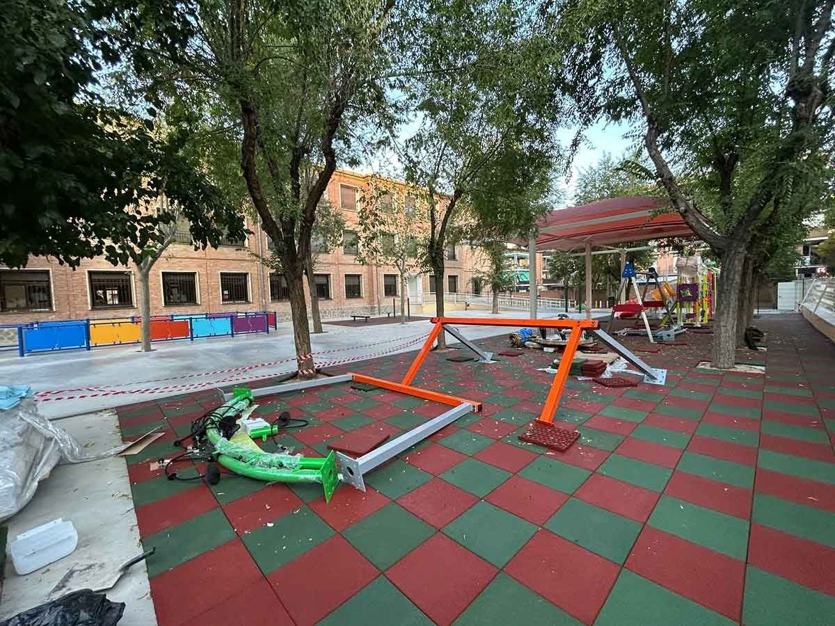 Parque infantil con baldosas de caucho verdes y rojas, un parque infantil naranja parcialmente desmontado y un edificio al fondo.