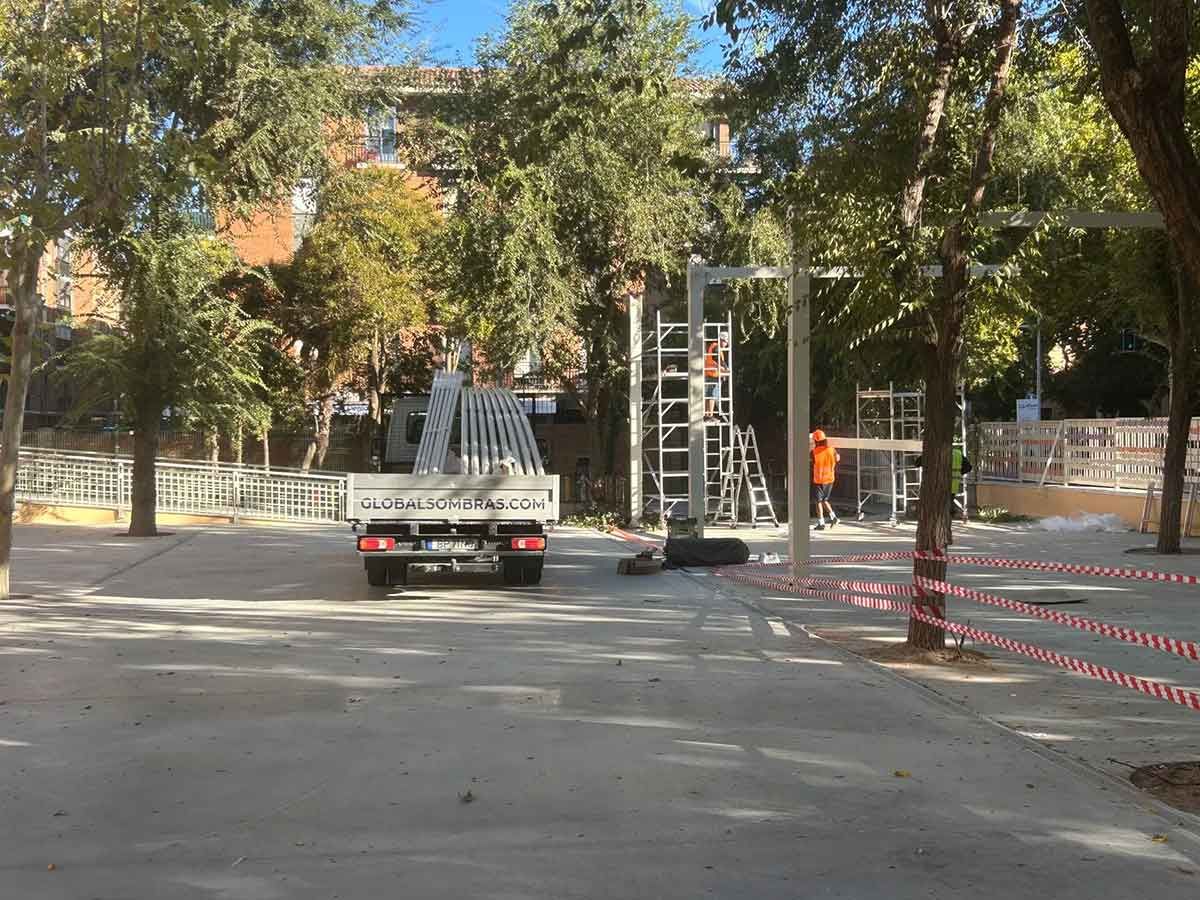 Camión estacionado en zona de construcción con andamios, árboles y un trabajador con chaleco naranja.