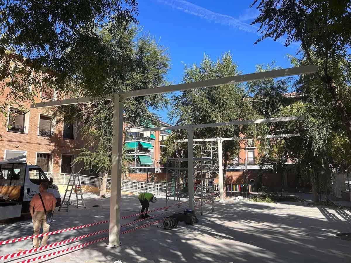 Un parque infantil en construcción con trabajadores, árboles y edificios al fondo. Estructura metálica blanca visible.