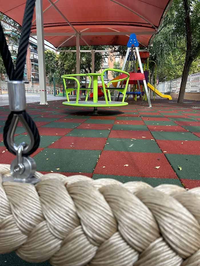 Escena de juegos: Cuerda de columpio en primer plano, con tiovivo y estructura de juegos bajo un dosel.