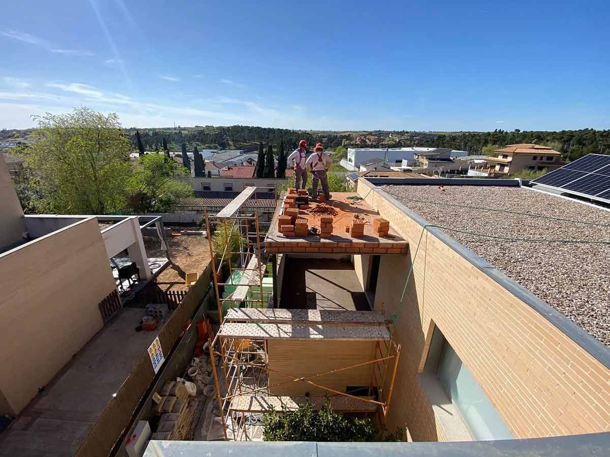 Dos trabajadores de la construcción en una azotea colocando ladrillos, día soleado con vista de la ciudad de fondo.