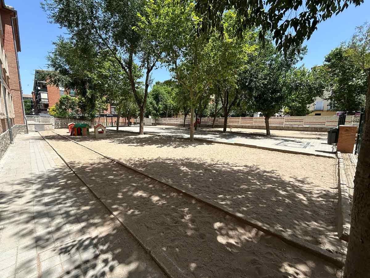 Parque de arena con árboles y una zona de juegos en un día soleado.