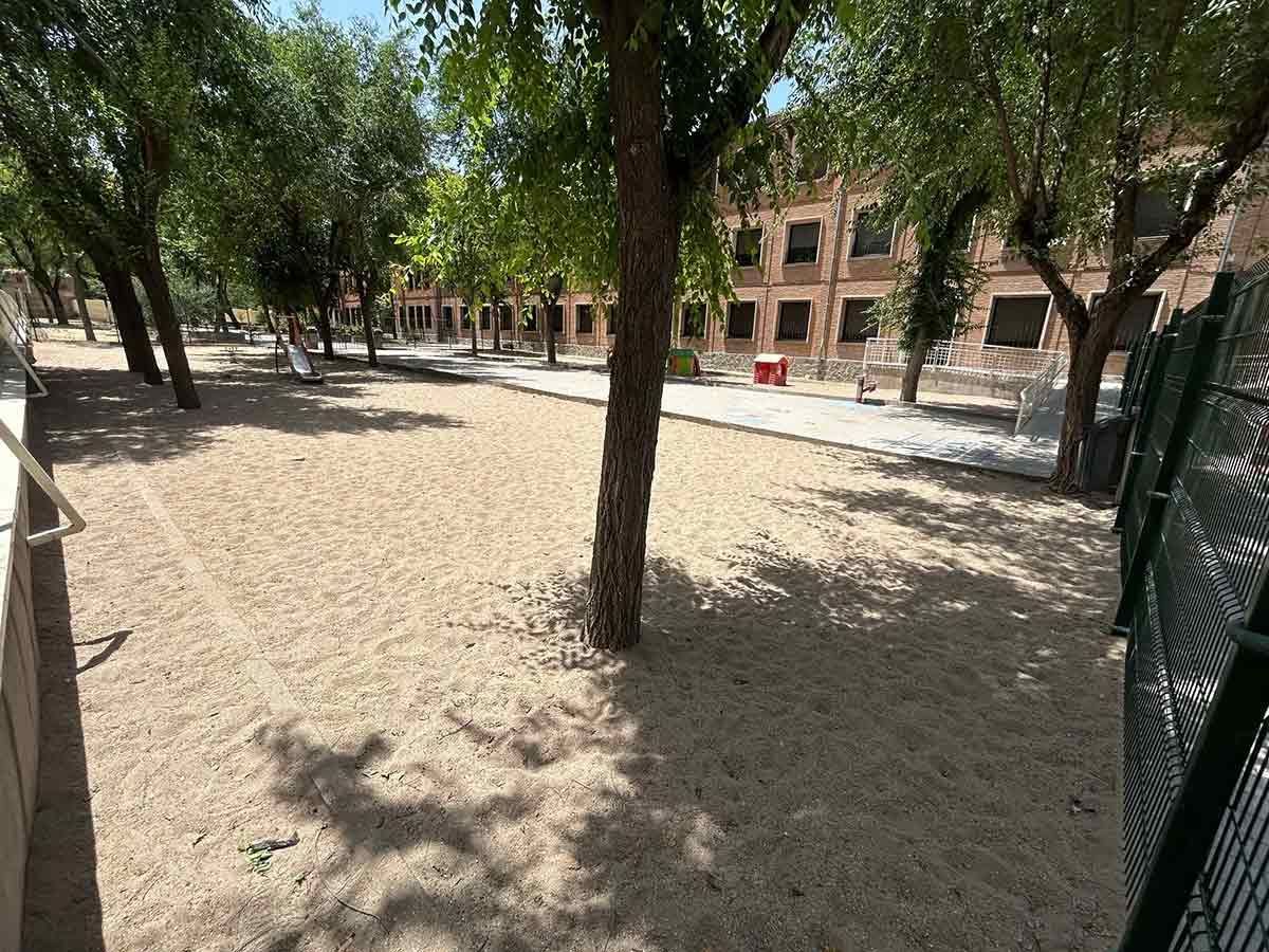 Patio de arena con árboles y un edificio al fondo, parcialmente cerrado por una valla verde.