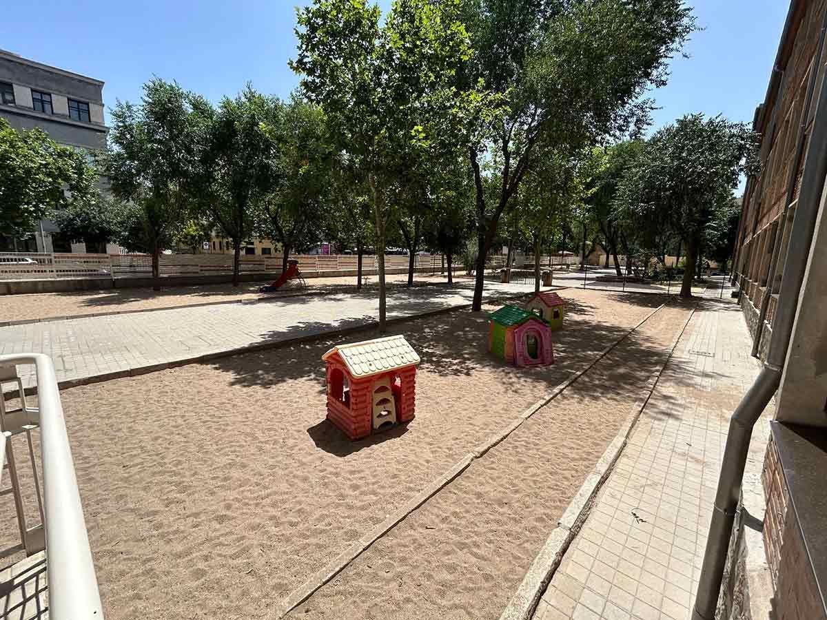 Parque infantil con casitas, arena, árboles y pasarela bajo un cielo soleado.