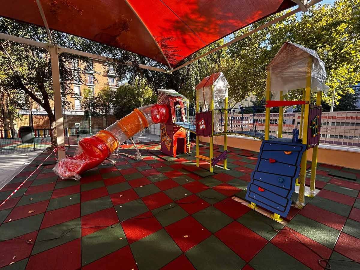 Parque infantil con tobogán rojo y naranja, muro de escalada y estructura de sombra, con suelo rojo y verde.