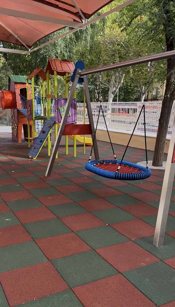 Parque infantil con columpios, tobogán y estructura de escalada bajo un dosel. Suelo de caucho rojo y verde.