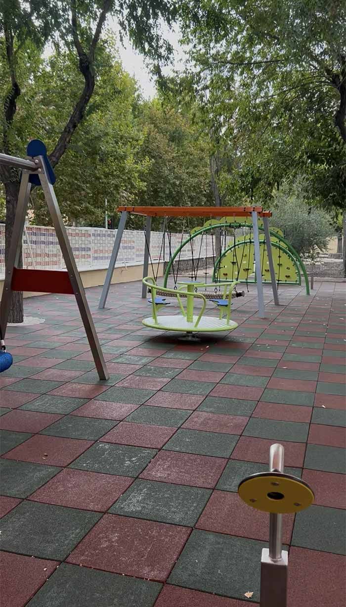 Parque infantil con columpios, tiovivo y estructura de escalada sobre superficie de caucho, rodeado de árboles.