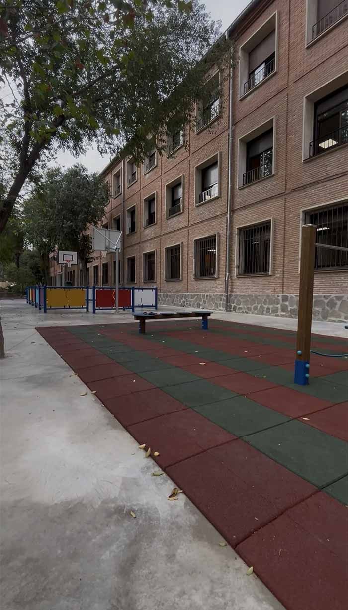 Patio de escuela con edificio de ladrillos, aro de baloncesto y suelo de caucho multicolor.