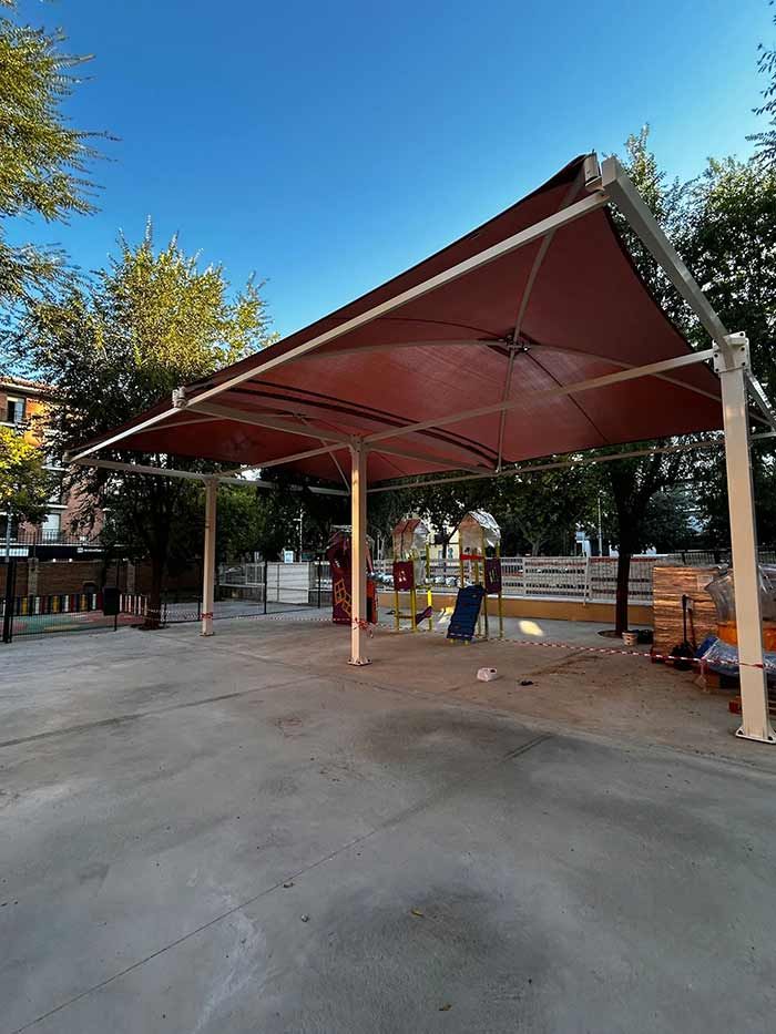 Toldo marrón sobre una zona de juegos de hormigón. Postes de soporte blancos, cielo azul y árboles al fondo.