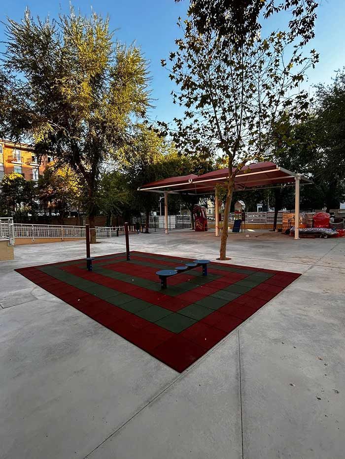 Parque con árboles, área de ejercicio con baldosas de caucho rojo/verde y una estructura sombreada.