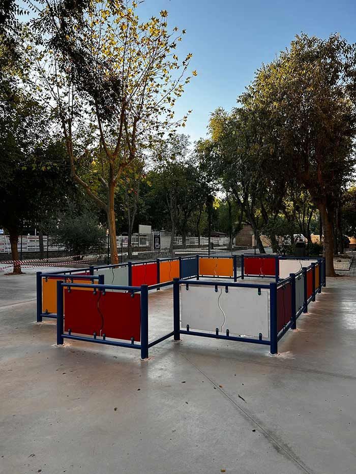 Una colorida estructura de laberinto con marcos azules y paneles de colores variados, en un entorno de parque con árboles.