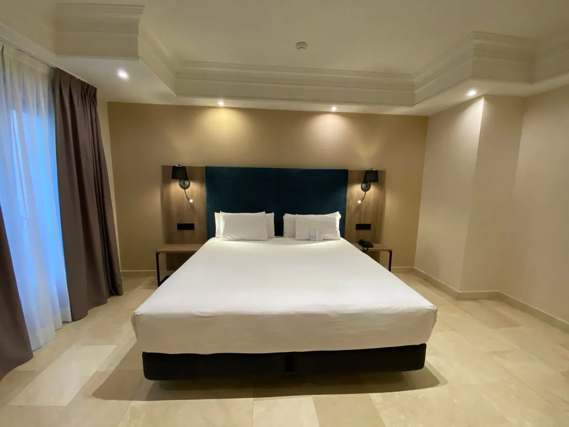 Dormitorio de hotel con cama tamaño king, cabecera color verde azulado, mesitas de noche y paredes de colores claros.