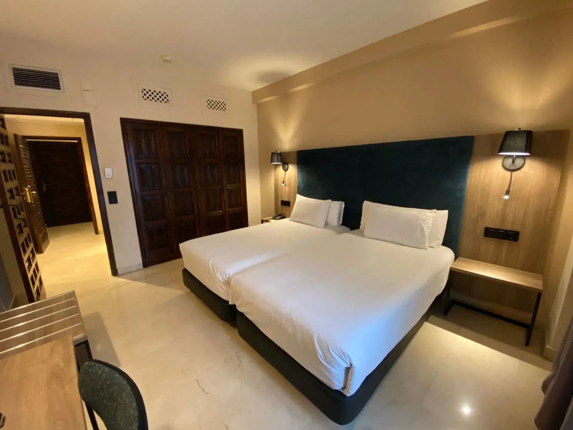 Dormitorio con dos camas individuales, cabecero oscuro, armario de madera, mesitas de noche y puerta de entrada.