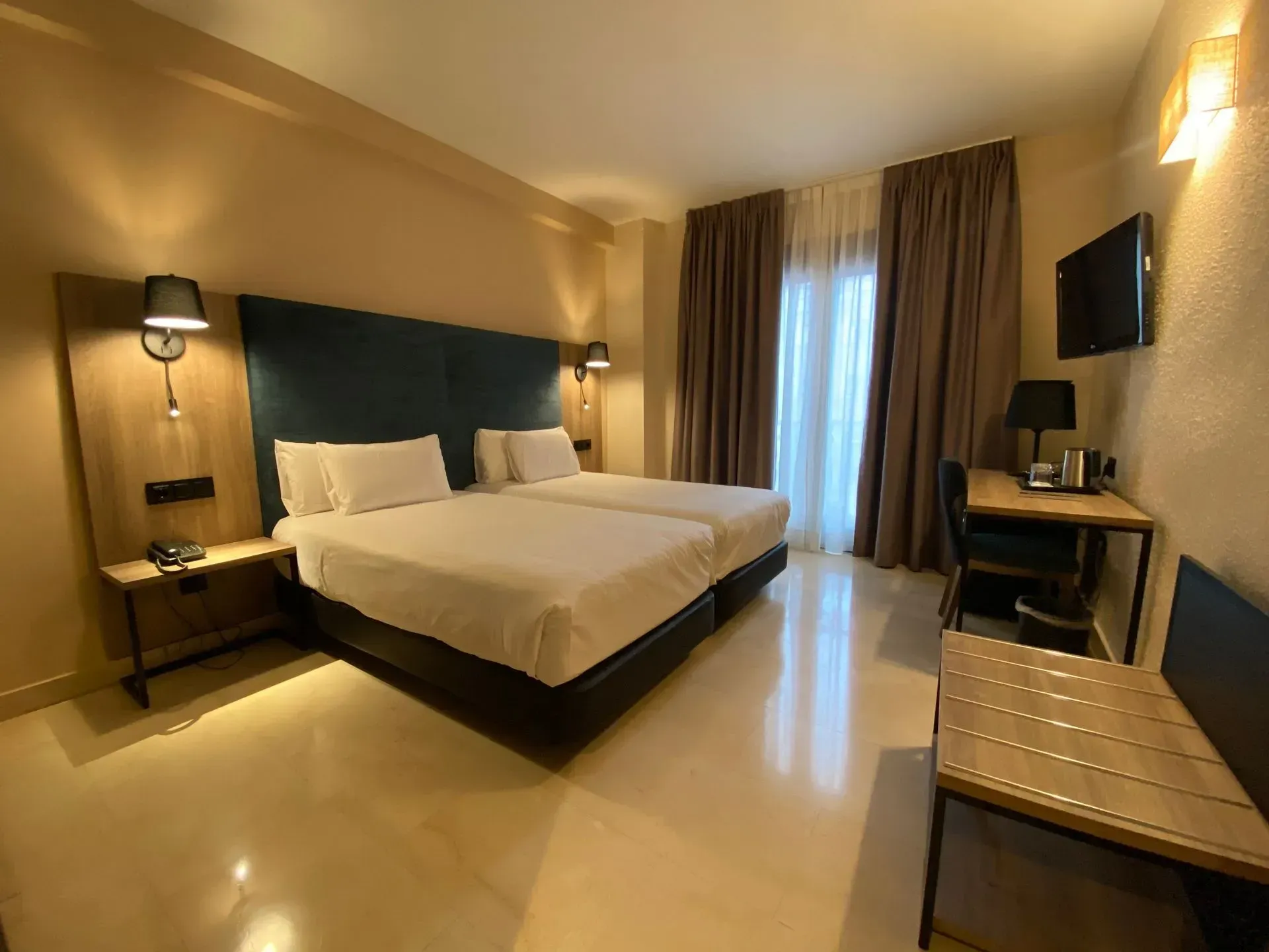 Habitación de hotel con dos camas individuales, un pequeño escritorio y una ventana con cortinas marrones.