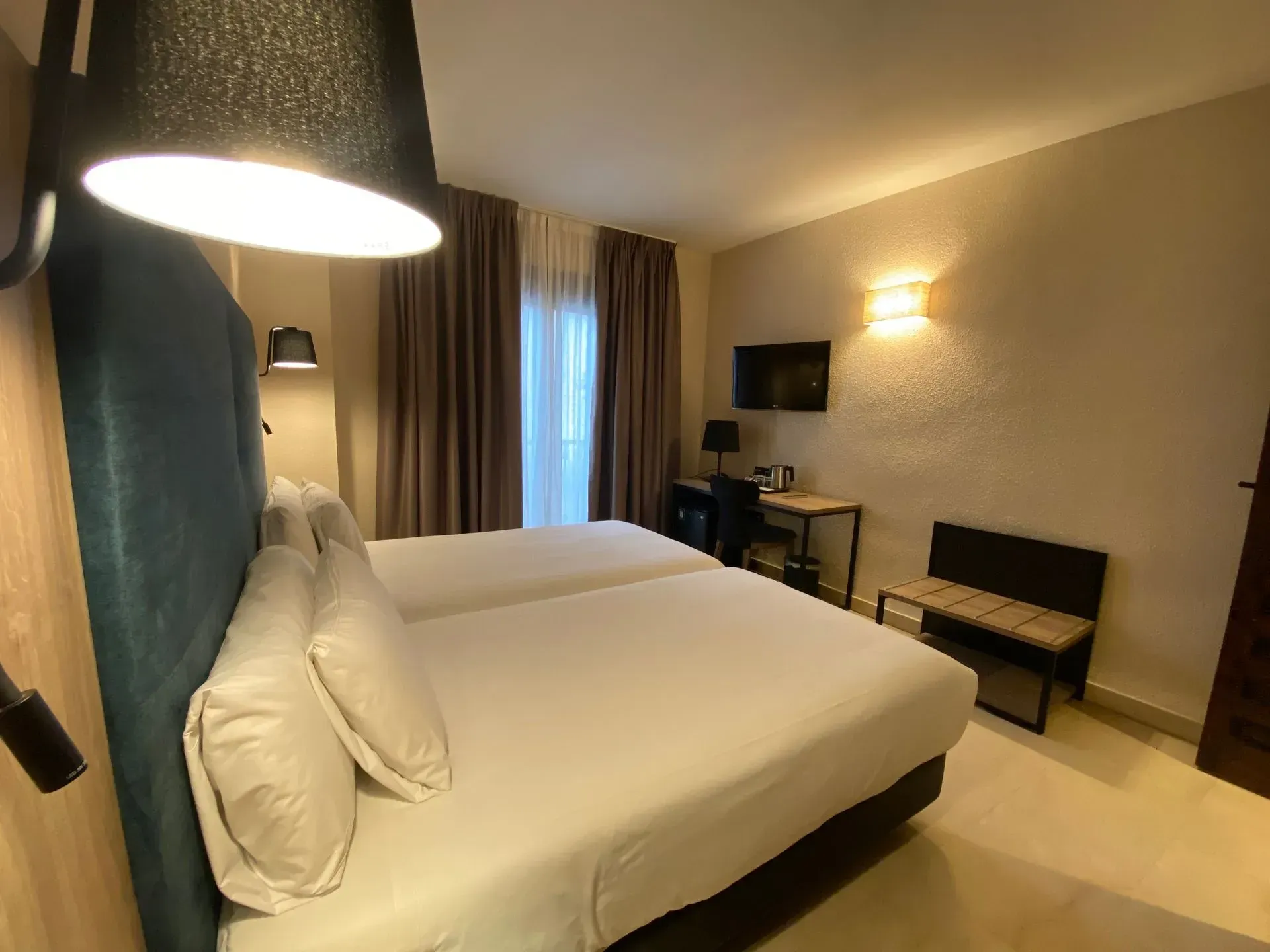 Habitación de hotel con dos camas, escritorio, televisor y ventana con cortinas. Iluminación suave y colores neutros.