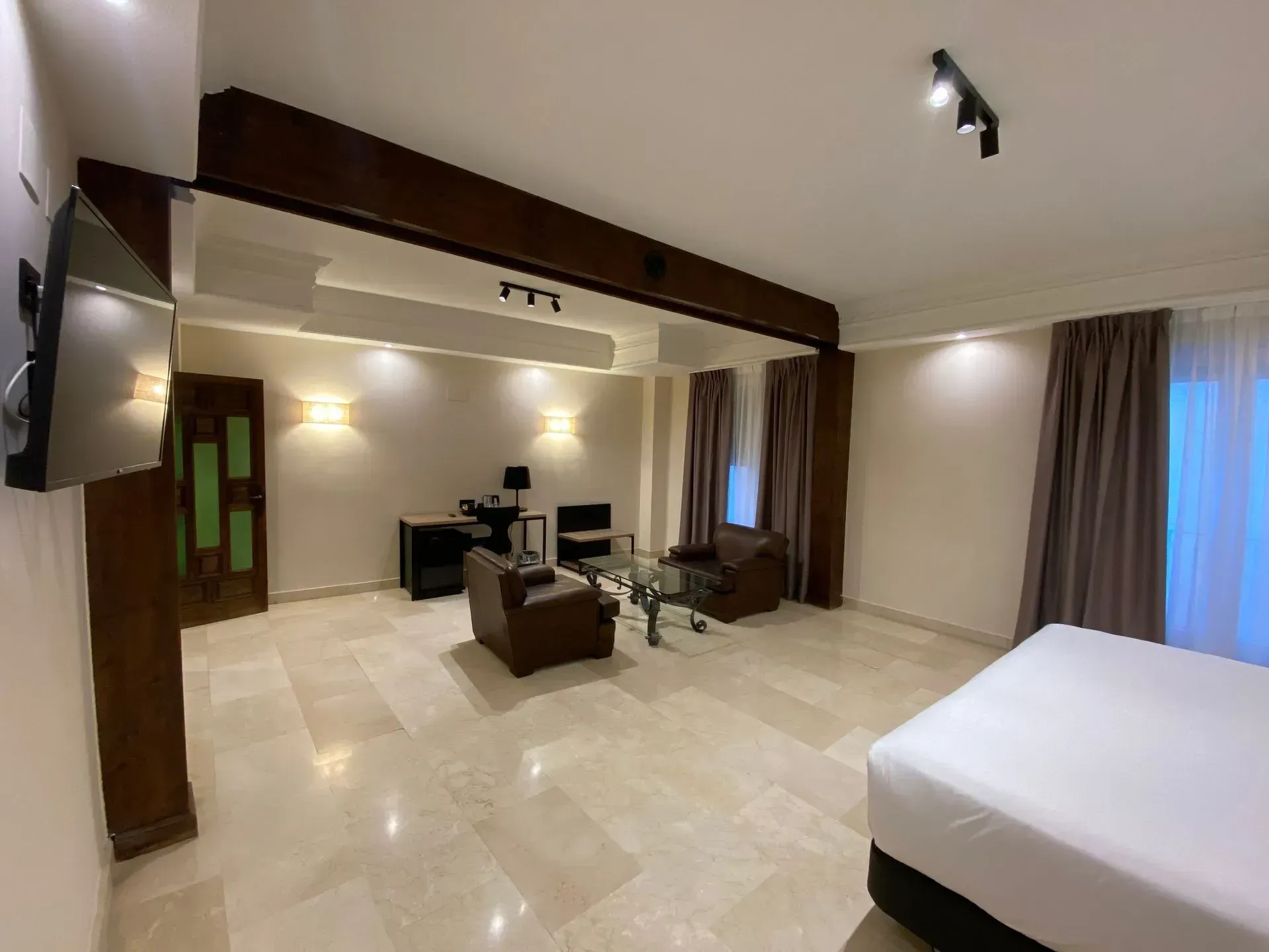 Amplia habitación de hotel con cama, zona de estar, escritorio y gran ventanal con cortinas.