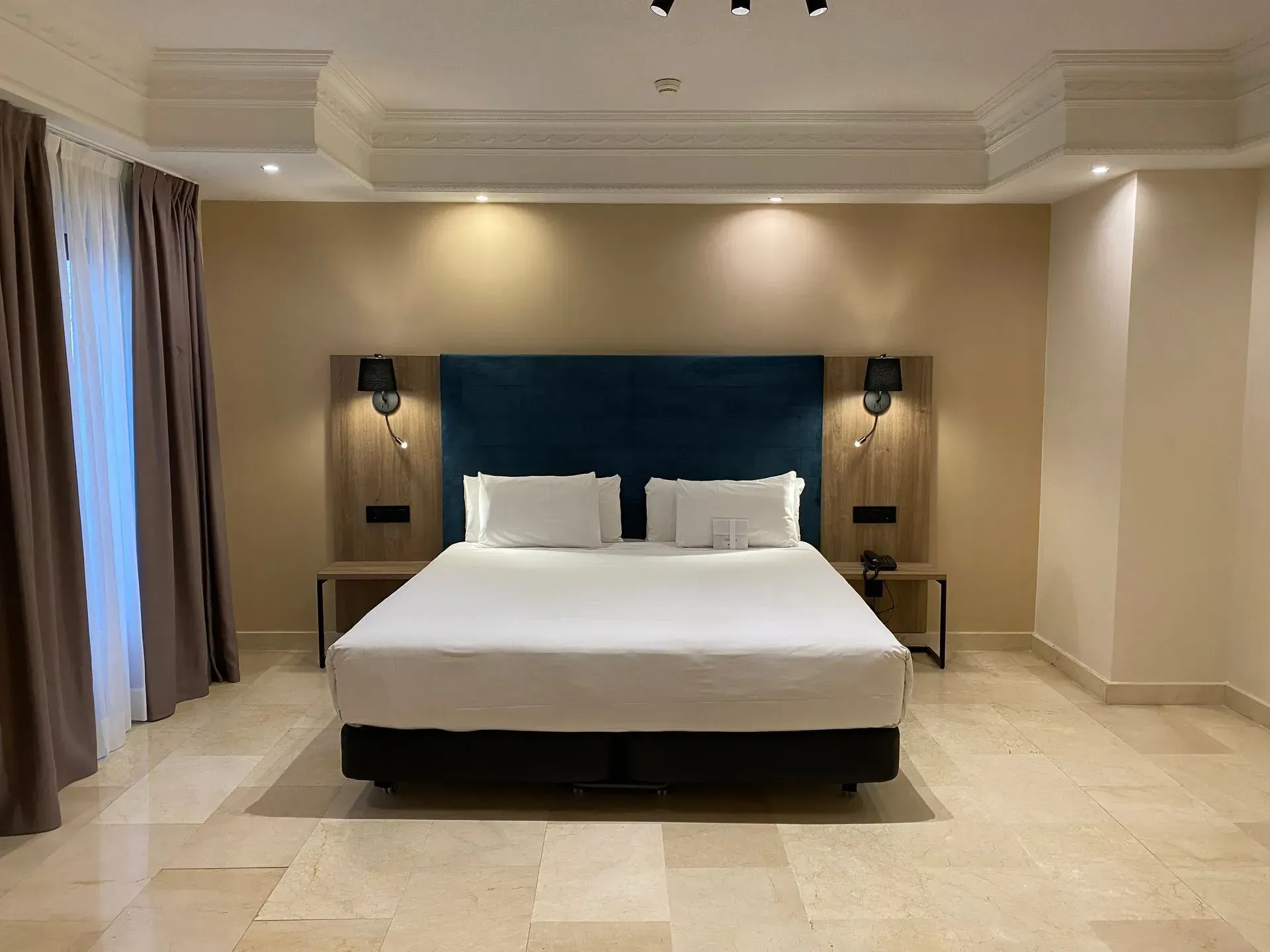 Dormitorio de hotel con cama tamaño king, cabecera color verde azulado, mesas auxiliares de madera, lámparas y paredes y suelos de color beige.