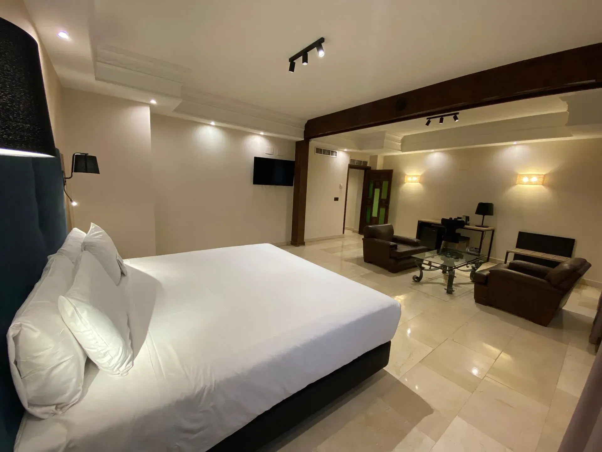 Habitación de hotel con una cama grande, zona de estar, TV y suelo de baldosas de color crema.