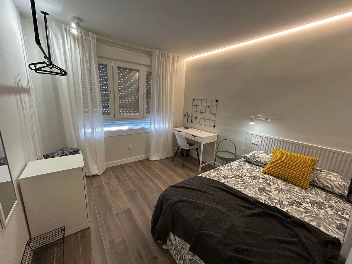 Interior de dormitorio con cama, escritorio junto a la ventana, cortinas blancas y una tira de luz.