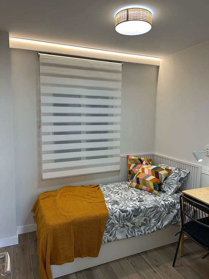 Dormitorio con cama, persianas y cojines decorativos. Una manta amarilla cubre la cama.