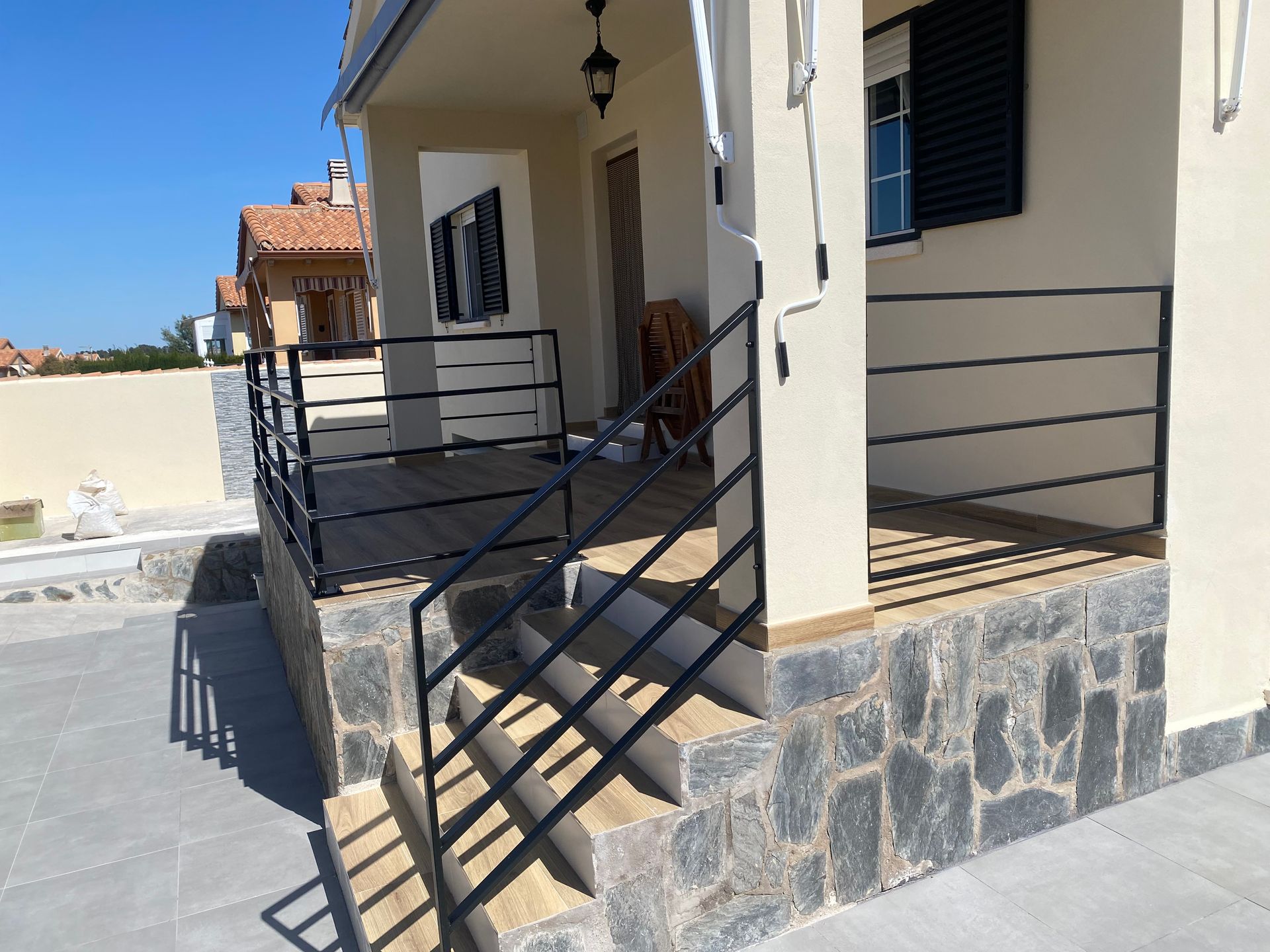 Exterior de una casa con porche y escaleras, con barandillas de metal negro y fachada de piedra, bajo un cielo despejado.