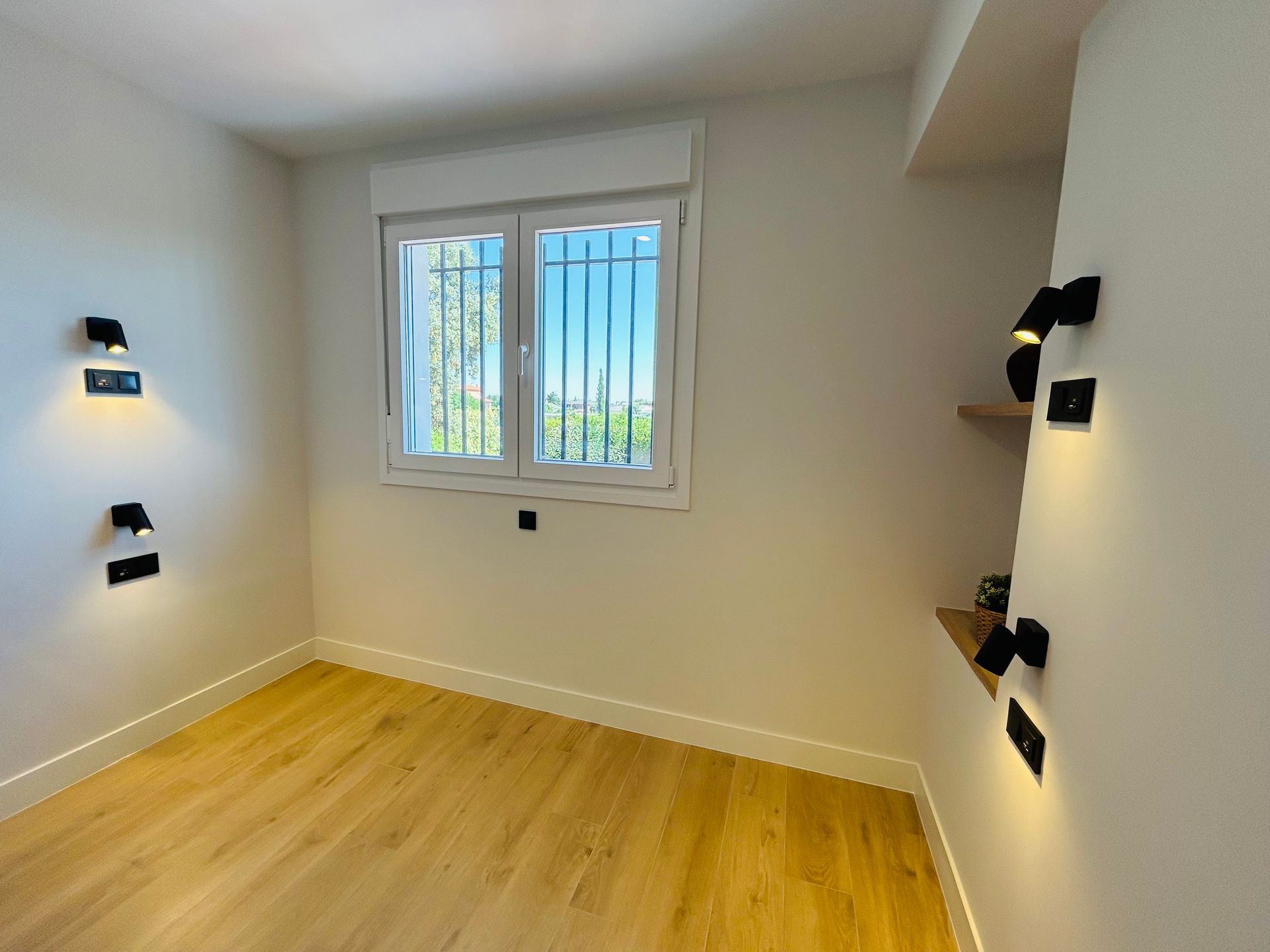 Habitación vacía con piso de madera, paredes blancas, ventana pequeña, apliques de pared negros y estantes.