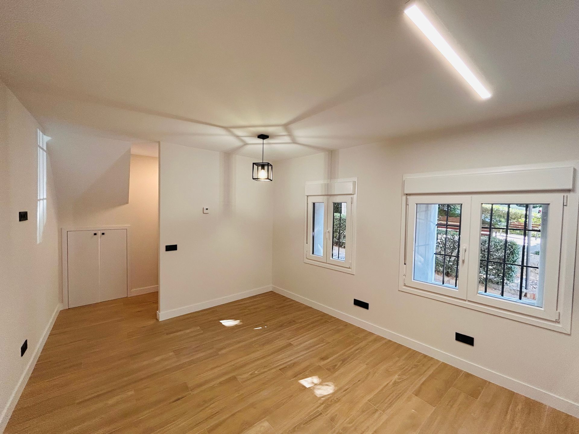 Habitación vacía con pisos de madera, dos ventanas e iluminación cenital.