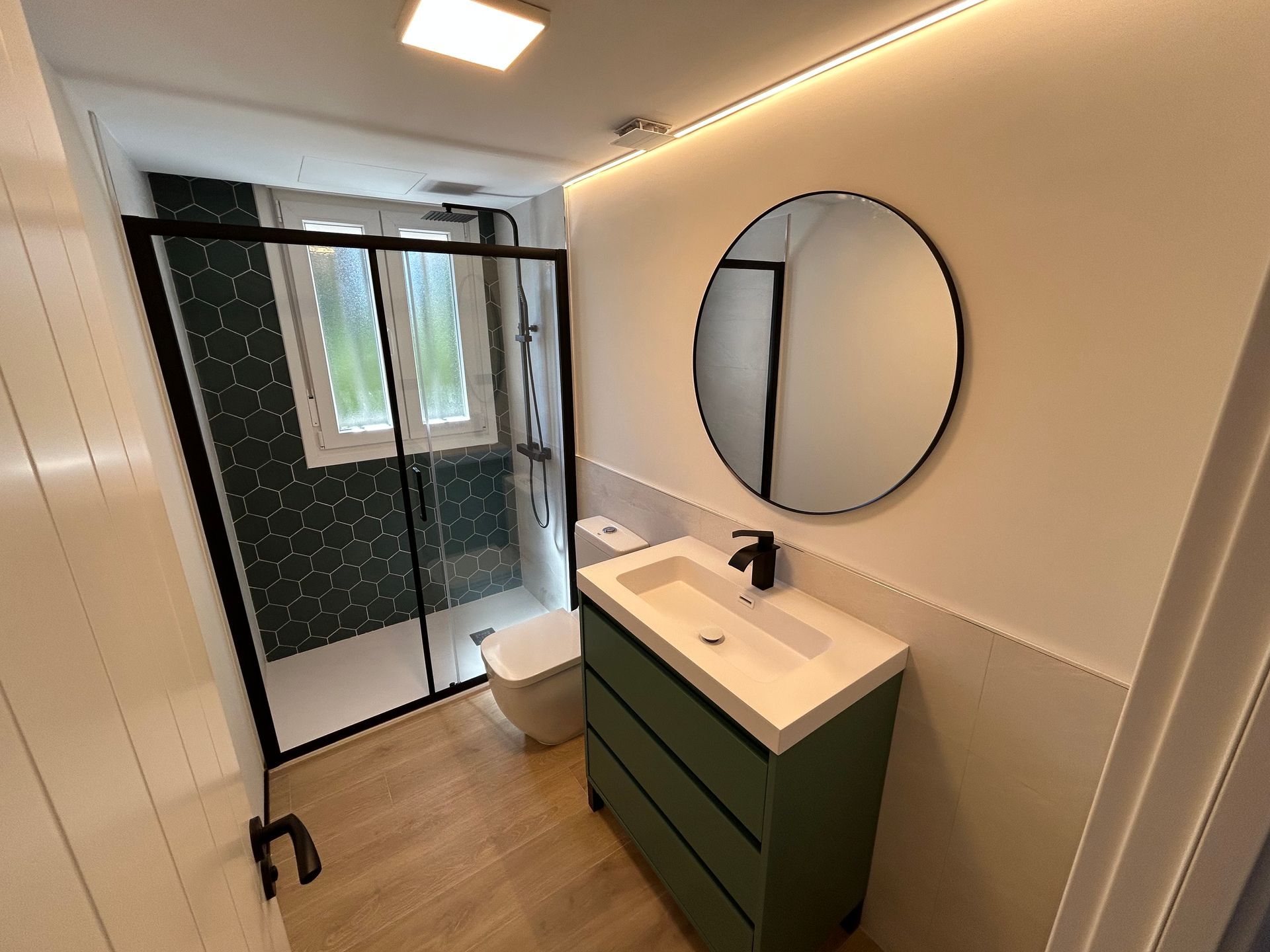 Baño moderno con tocador verde, espejo redondo y ducha con marco negro.