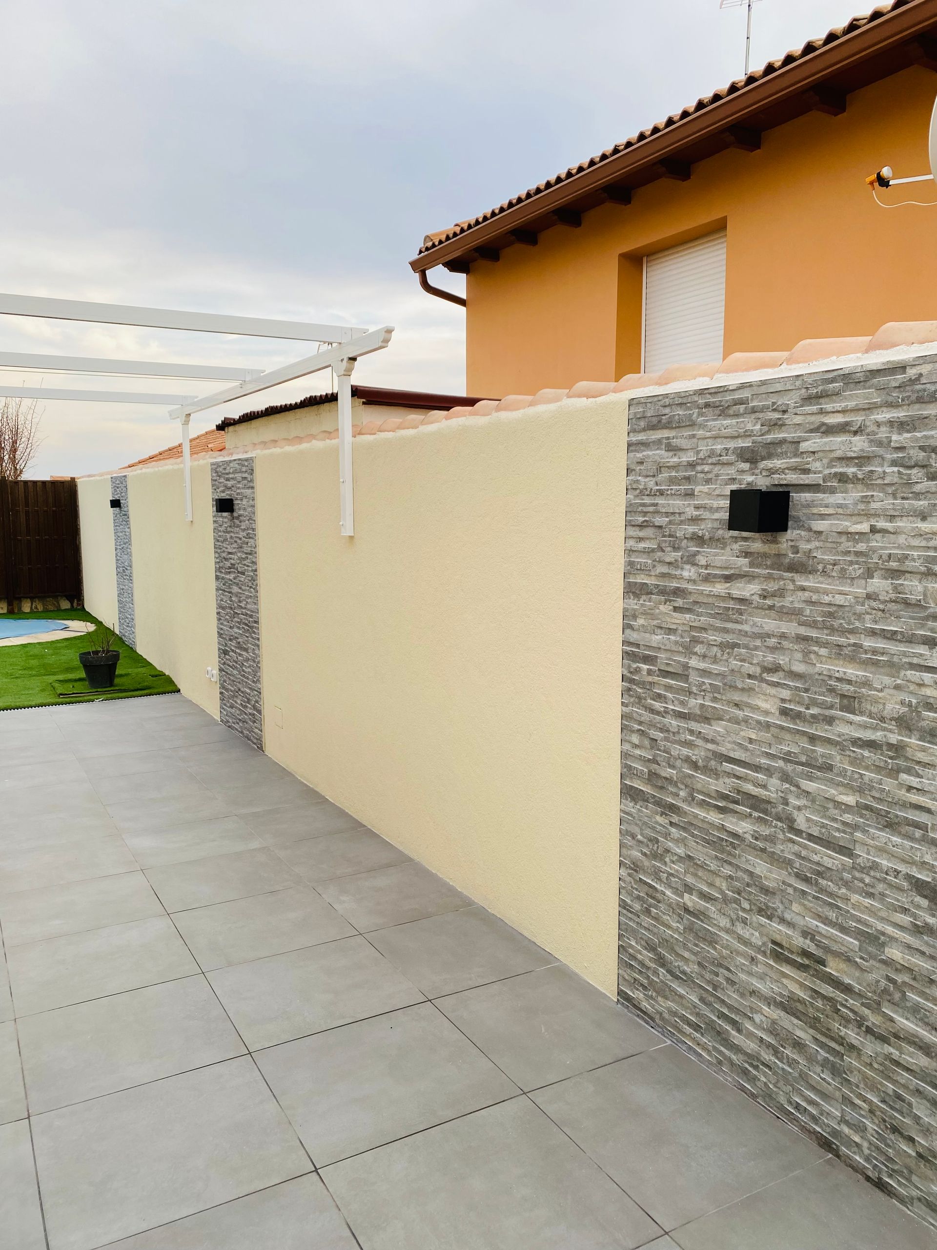 Muro largo con secciones grises y beige, detalles de piedra, iluminación moderna y patio embaldosado.